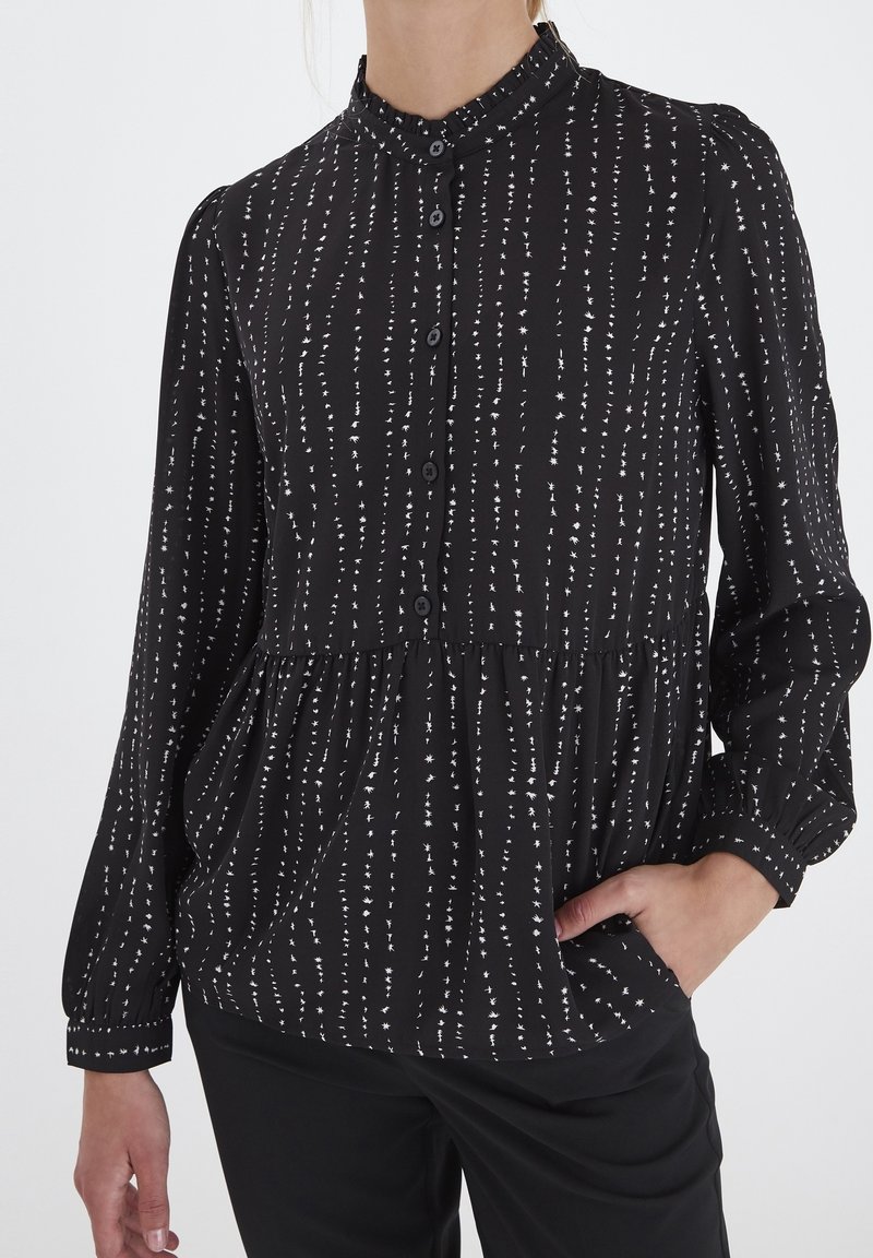 Blouse noire avec des motifs d'étoiles blanches verticales, manches longues, devant boutonné et taille légèrement froncée portée par une personne avec une main dans la poche.