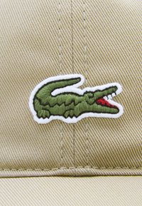 Kačket u boji kaki s zelenim izvezenim logotipom krokodila s crvenim naglascima, postavljen na teksturiranu tkaninu u pozadini.