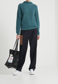 Maglione a coste teal con collo alto, abbinato a pantaloni blu navy con una tasca a toppa marrone e una borsa tote nera con dettagli bianchi.