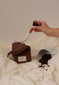 Porte-pinceaux cosmétique marron avec une fermeture éclair à accents dorés, contenant des pinceaux. Pinceau à manche blanc dans la main, grains de café éparpillés, et mug blanc.