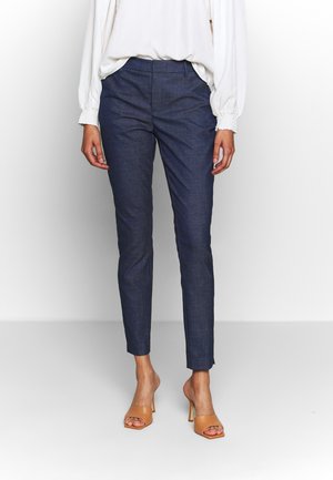 Woman wearing dark blue slim-fit trousers, white long-sleeve blouse, and tan open-toe heeled sandals, standing against a plain background.