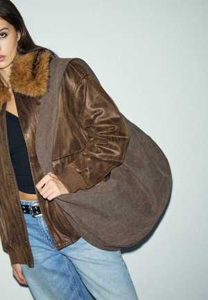 Donna con una giacca di pelle marrone con colletto di pelliccia, top nero, jeans blu e che porta una grande borsa a tracolla marrone.