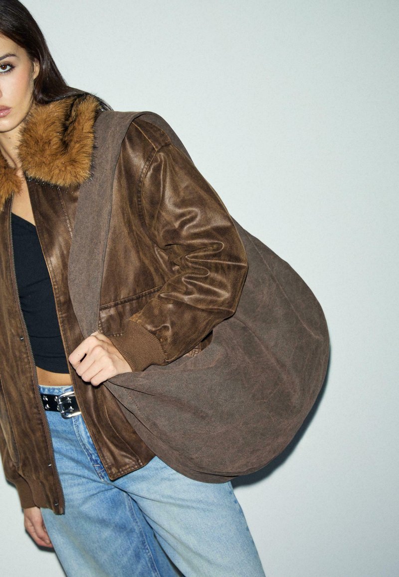 Kvinde iført en brun læderjakke med pelskrave, sort top, blå jeans og bærende på en stor brun skuldertaske.