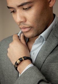 Black braided bracelet with gold accents worn on the wrist. The individual is in a gray suit with a light blue shirt, adjusting the collar.