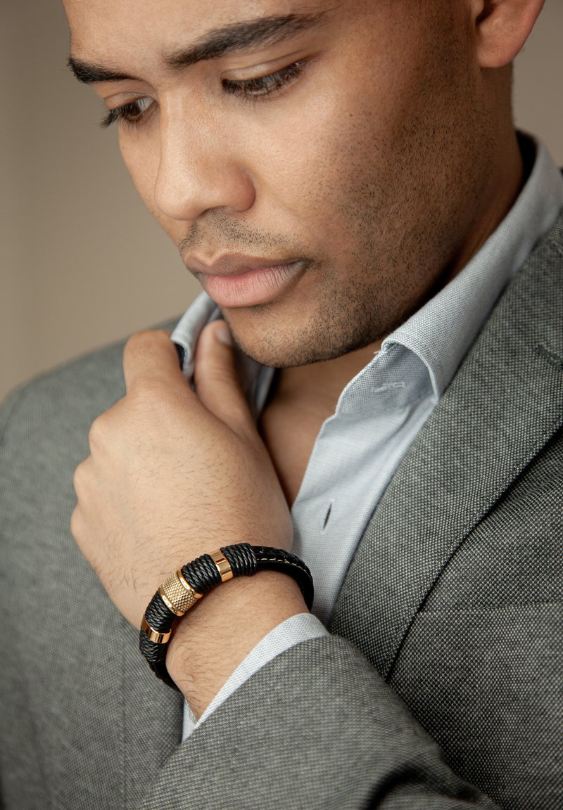 Black braided bracelet with gold accents worn on the wrist. The individual is in a gray suit with a light blue shirt, adjusting the collar.