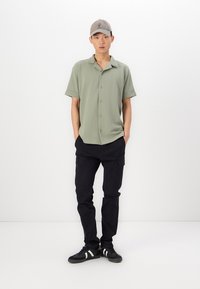 Kortärmad, ljusgrön polo-tröja med knappar, ihop med svarta cargobyxor. Modellen har på sig en ljusgrå keps och svarta sneakers.