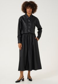 Melna mākslīgās ādas vidēja garuma jaka ar pogām un divām priekšējām kabatām, kombinēta ar plūstošu melnu midi svārku. Melni papēžu kurpes noslēdz izskatu.