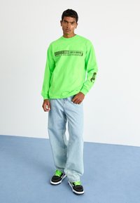 Neonově zelená košile s dlouhým rukávem obsahuje grafický text, doplněná o světle modré volné džíny a černé tenisky se zelenými tkaničkami.