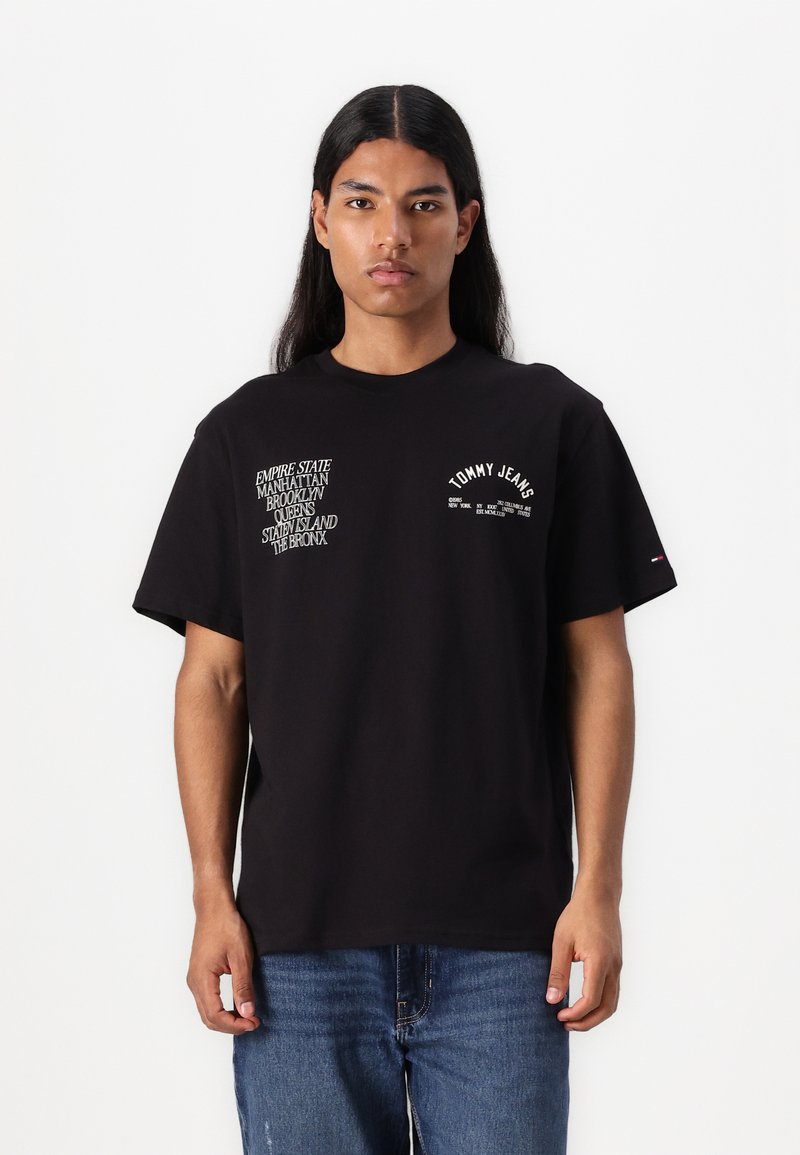 Camiseta de algodón negra con gráficos de texto blanco en el lado izquierdo y derecho del pecho; ajuste relajado y cuello redondo. Se lleva con jeans azules.