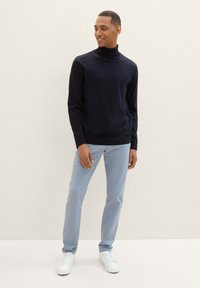 Navy turtleneck sweater, ribbed texture, paired with light blue trousers and white sneakers. Model stands casually against a plain background.