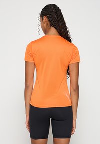 Pomarańczowy sportowy t-shirt z krótkim rękawem, teksturowaną tkaniną i siateczkowymi wstawkami przy krawędzi. W zestawie z czarnymi szortami.