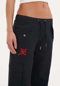 Zwarte cargobroek met een trekkoord in de taille, zijzakken en een rood geborduurd logo. Lichtgewicht stof met een gladde textuur.
