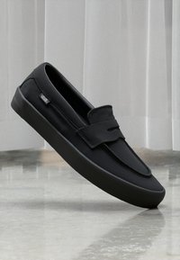 Sorte lerret slip-on sko med rund tå, frontrem-detalj og gummisåle. Merket "Vans" på siden. Glatt tekstur.