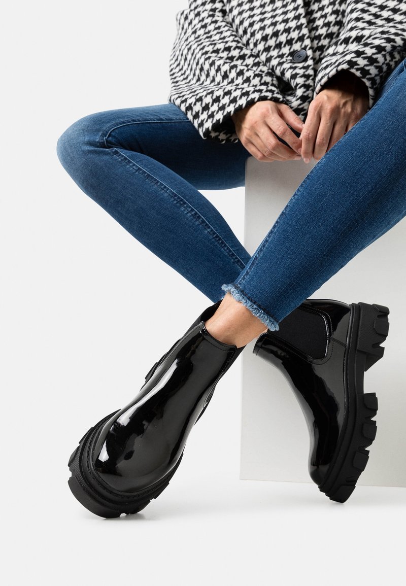 Personne assise portant un jean skinny bleu, une veste pied-de-poule noire et blanche, et des bottines noires chunky brillantes.