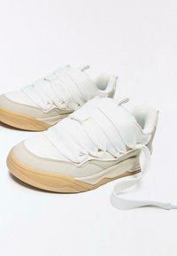 Fehér tornacipő gumitalppal, texturált, párnázott felsőrésszel és egyedi pántdizájnnal. Egyszínű, lágy részletekkel.