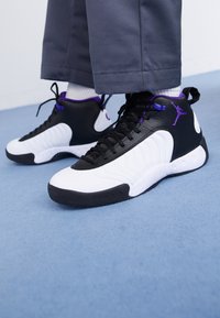 Osoba oblečená v černých, bílých a fialových basketbalových teniskách Air Jordan s černými tkaničkami a šedých kalhotách na modrém koberci.