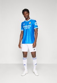 Blue football jersey with white accents, featuring the brand logo and team name. Worn with white shorts, blue-striped socks, and football shoes.