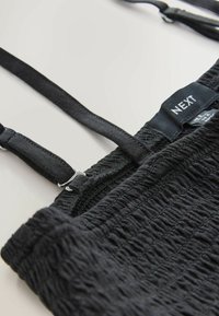 Material texturat negru cu detalii pliate, cu bretele reglabile și o etichetă inscripționată cu „NEXT”. Suprafață netedă cu modele subtile.