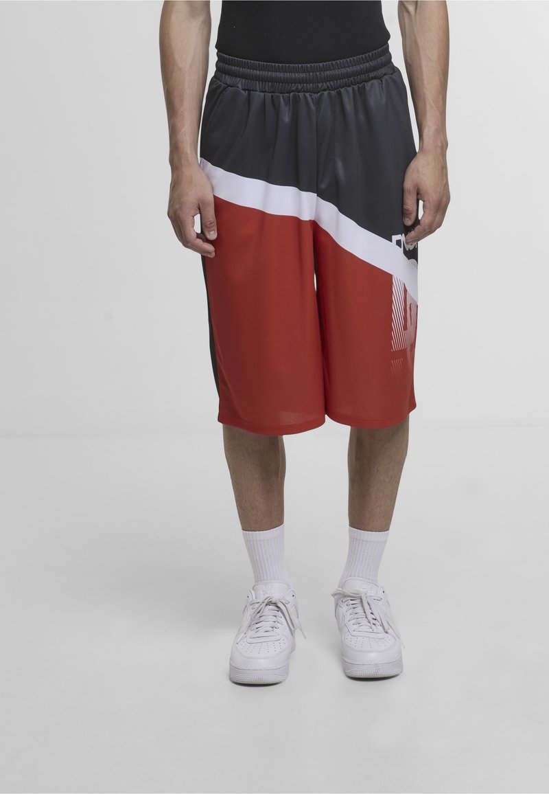 Homme portant un short de basket en bloc de couleurs noir, blanc et rouge, des baskets blanches et des chaussettes blanches montantes, debout sur un fond clair uni.