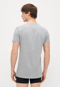 Γκρι κοντομάνικο μπλουζάκι από βαμβάκι με ελαφρώς στρογγυλή λαιμόκοψη και ίσια ανοιχτή. Φοράει σκούρα μπλε μποξεράκια.