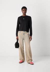 Schwarzes Langarmshirt mit einem türkisfarbenen CK-Logo, lockere helle Denim-Jeans und pinke Sneakers, hält eine kleine schwarze Tasche.
