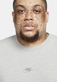 Hallitatud hall ringkaelusega särk, millel on tikitud Nike logo. Nahk näitab silmapaistvaid freckles ja habe. Lihtne disain, pehme tekstuur.
