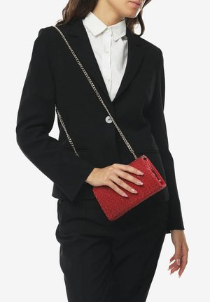 Femme portant un costume noir et une chemise blanche, tenant un petit sac bandoulière rouge texturé avec une chaîne argentée.
