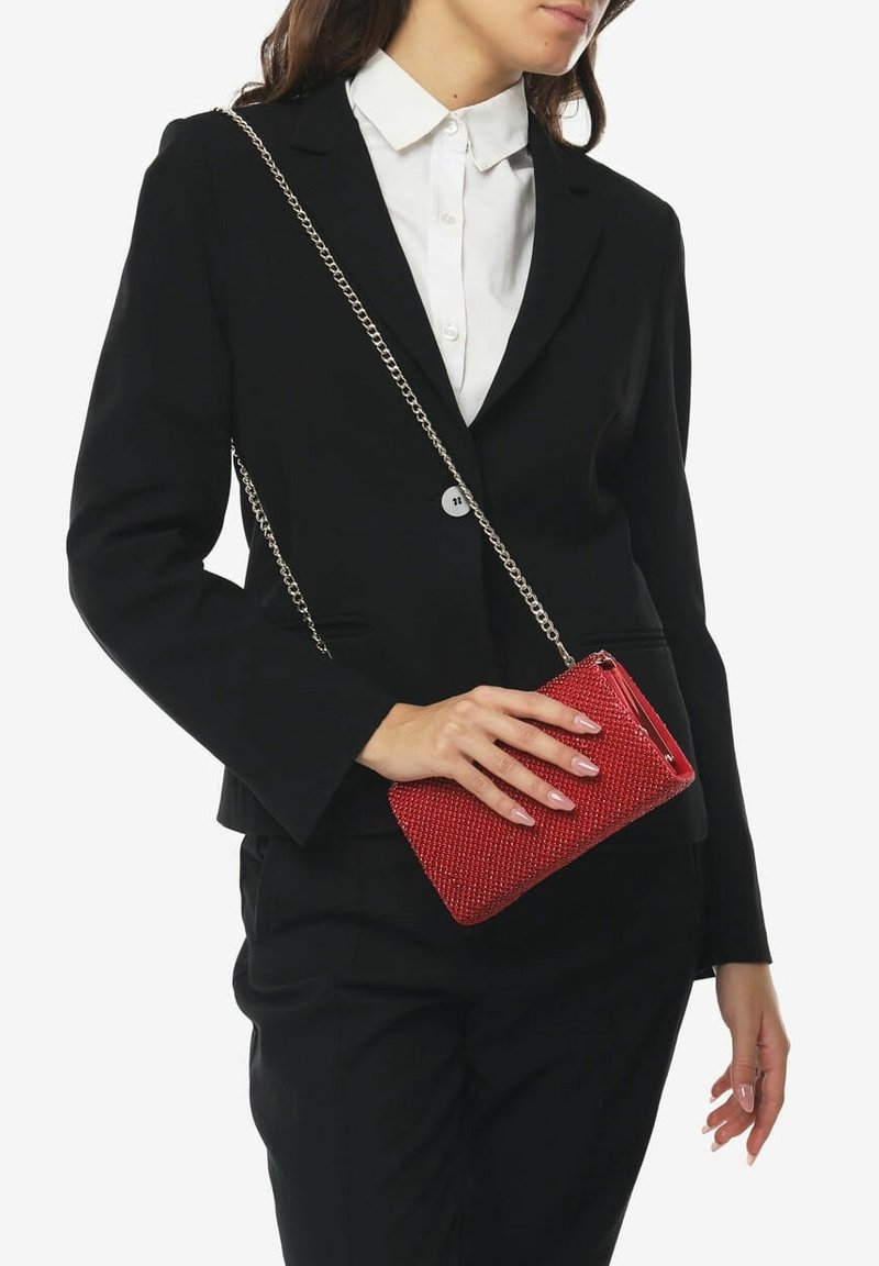 Femme portant un costume noir et une chemise blanche, tenant un petit sac bandoulière rouge texturé avec une chaîne argentée.