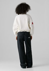 Πουλόβερ από λευκό φλις μεγάλου μεγέθους με σούρες στη μέση, συνδυασμένο με άνετα μαύρα τζιν με κόκκινες ραφές και λεπτομέρειες με λογότυπο στο πίσω μέρος.