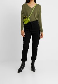 Maglia verde oliva a maniche lunghe con scollo a V, pantaloni cargo neri con tasche e stivali neri alla caviglia con punta appuntita; piccola borsa a tracolla verde.