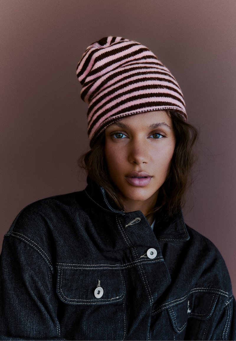 Jeune personne à la peau claire et aux yeux bleus portant un bonnet rayé rose et noir et une veste en jean foncé devant un fond uni.
