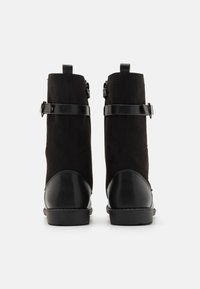 Bottes noires à la cheville en daim avec des accents en cuir. Elles présentent une sangle avec une boucle et un bout arrondi. Semelle plate.
