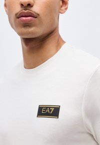 Nuori mies, joka pukee valkoisen pyöreäkauluksisen paidan, jossa on musta ja kultainen EA7 Emporio Armani -logo rintapohjassa.