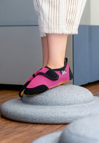 Schwarze und rosa Netzschuhe mit Klettverschlüssen und einer flexiblen Sohle, auf einem runden grauen Kissen auf einem Holzfußboden platziert.