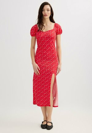 Mujer con vestido midi rojo floral con mangas abullonadas y abertura lateral, combinado con zapatos Mary Jane negros, de pie frente a un fondo liso.