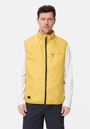 Man die een gele mouwloze gilet met ritssluiting draagt over een wit shirt met lange mouwen en donkere korte broek, staand tegen een effen achtergrond.