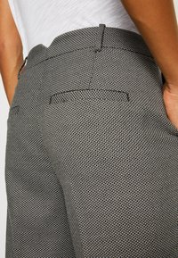 Gros plan d'une personne portant un pantalon à motifs noirs et blancs avec des poches passepoilées à l'arrière et un haut blanc rentré.