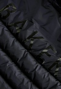 Marineblå pufferjakke med en quiltet design og glinsende sort bogstaver "BAKER" hen over fronten, der fremhæver en glat tekstur.