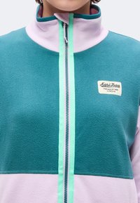 Γυναίκα που φοράει fleece μπουφάν με πράσινα-μπλε και ανοιχτό μοβ πάνελ και φερμουάρ μπροστά, εμφανίζοντας ένα μπάλωμα με το κείμενο "Kari Traa."