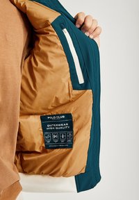 Teal ytterjacka med en beige isolerad foder, med kviltad textur, en vit dragkedjeficka och en blå skötselguide med ikoner.