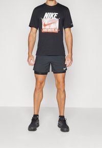 Černé tričko Nike s grafickým designem a nápisem "JUST DO IT" v bílé a červené barvě, spárované s černými šortkami a černými sportovními botami.