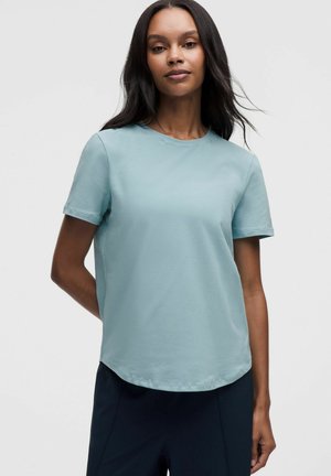 Donna con capelli lunghi e scuri che indossa una t-shirt azzurro chiaro a maniche corte e pantaloni scuri su uno sfondo chiaro e semplice.