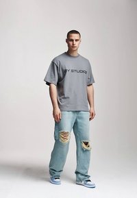 Siva oversize majica sa tekstom "BY STUDIOS", uparena s blijedoplavim trapericama sa velikim poderotinama. Izgled upotpunjuju tenisice.