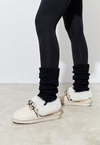 Pantofola beige con bordi in pelliccia sintetica bianca, dettagli neri e logo. Abbinata a gambaletti neri a coste e leggings neri.