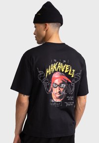 Svart bomull t-shirt med grafisk tryck av ett ansikte med en röd bandana. Texten inkluderar "MAKAVELI" och "THUG LIFE" i starka färger.