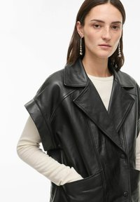 Trench-coat sans manches en cuir noir avec un col cranté, de grandes poches avant et une fermeture à boutons. Porté par-dessus un haut à manches longues de couleur claire.