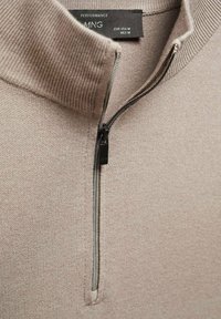 Pull en tricot beige avec une fermeture éclair noire à moitié ouverte et un col montant côtelé, étiquette MNG Performance, taille moyenne.