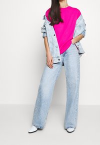 T-shirt rose vif, porté sous une veste en denim délavé, associé à un jean ample bleu clair et des bottes blanches à bout pointu.