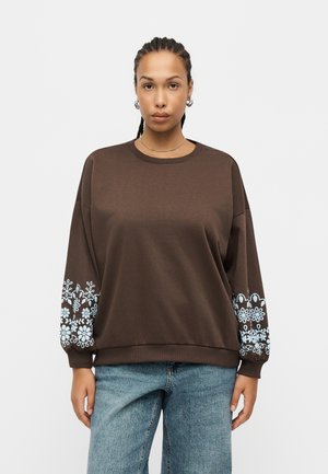 Frau blickt nach vorne, trägt ein braunes Sweatshirt mit hellblauen Blumenmustern an den Ärmeln und blaue Jeans, vor einem einfarbigen Hintergrund.
