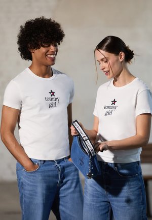 Fehér pólók "tommy girl" logóval és csillaggal. Világoskék farmernadrág. A nő egy kék farmer táskát tart, fekete pánttal és szív charm-mal.